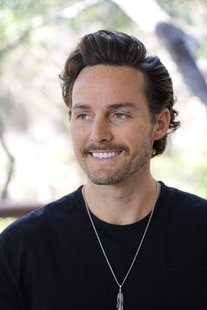 Allan Butler headshot, smiling outdoors with blurred trees in the background, wearing a black sweater and silver feather necklace